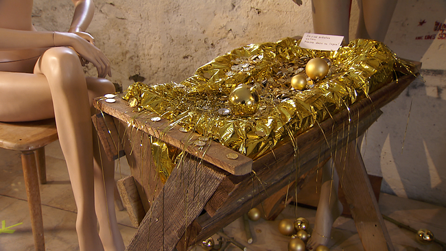 Holzkrippe mit goldenem Inhalt, am Rand sitzen und stehen 2 unbekleidete Schaufensterpuppen