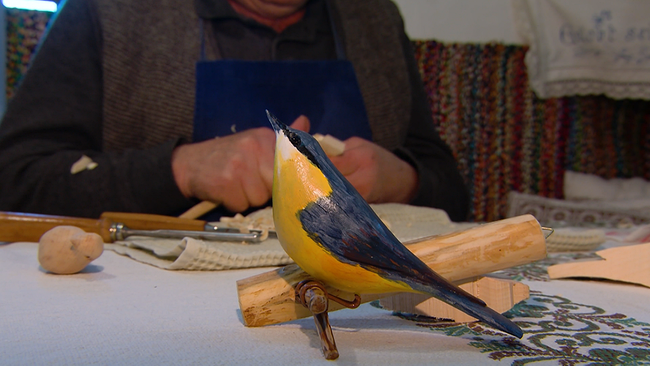 Im Vordergrund ein bemalter, aus Holz geschnitzer Vogel, im Hintergrund Hände, die gerade Holz schnitzen