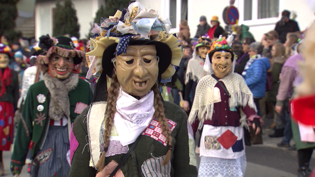 Mehrere Personen mit holzgeschnitzten Masken und selbstgemachten Kostümen bei einer Faschingsparade auf der Straße