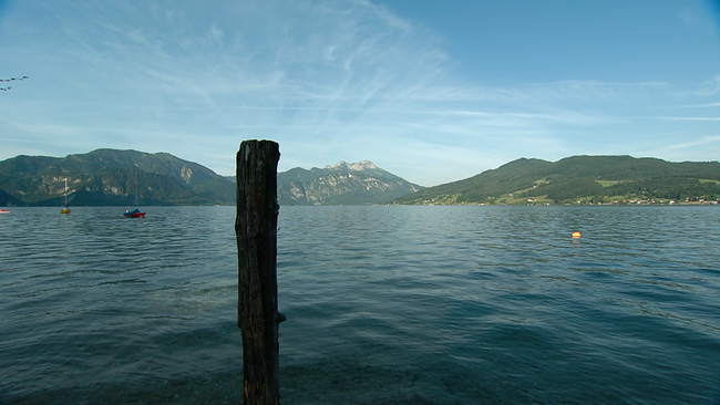 Aus dem Wasser, mitten in einem blauen See, ragt ein Holzpflock, an dem Boote festgemacht werden können. Im Hintergrund erheben sich sanfte grüne Hügel, die den See begrenzen