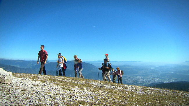 Eine Wandergruppe, die hintereinander auf einem Berggrad gehen. Im Hintergrund, strahlend blauer Himmel und die Bergketten der umliegenden Landerhebungen