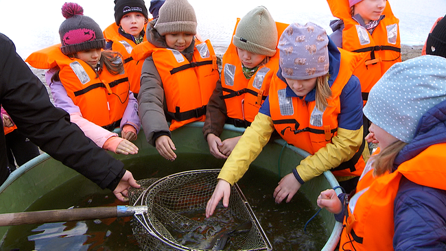 Kinder in Schwimmwesten stehen um großen Bottich herum und blicken auf Fisch im Netz