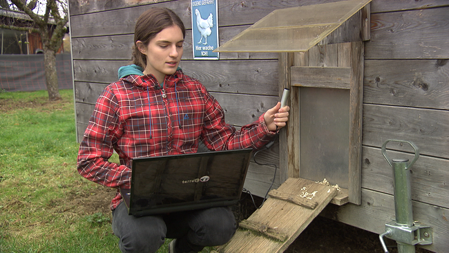 Junge Frau sitzt mit Laptop und Mikrofon vor einem Hühnerstall