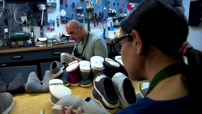 Zwei Personen, die an einem Arbeitstisch sitzen. Auf dem Tisch sind etwa 12 Paar Schuhe, die sowohl von der Person im Vordergrund, als auch von der im Hintergrund bearbeitet und bestickt werden.