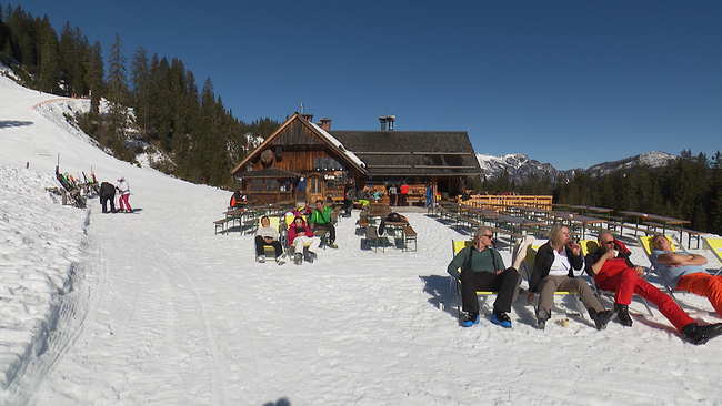Ein Panoramabild mit einer Hütte vor einem verschneiten Berg. Vor der Hütte sitzen mehrere Personen und genießen die Sonne. 