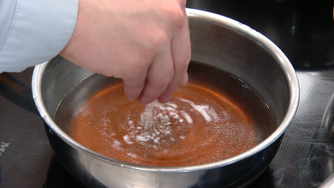 Eine Person, die zwischen ihren Fingern etwas Salz in einen Kessel mit Suppe rieseln lässt. In der klaren Brühe schmilzt das Salz sofort 
