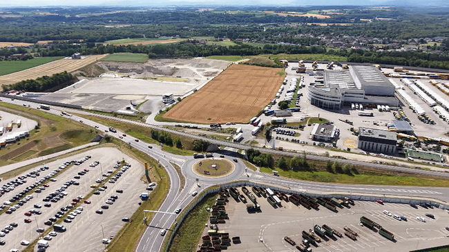 Ein Blick aus der Luft auf eine dicht verbaute Straße mit Kreisverkehr und Industrieanlagen an denen gebaut wird. Dazwischen, einsam, ein Acker. 