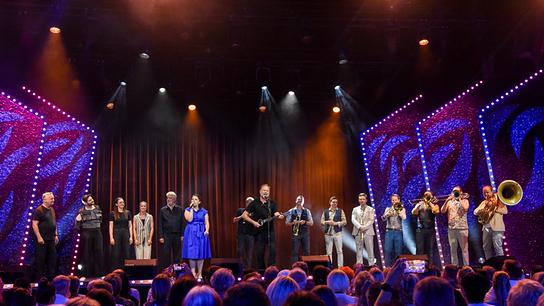 Thomas Stipsits steht mit Gitarre auf der Bühne von "Andreas Vitásek & Friends" begleitet von Isabell Pannagl, Christof Spörk und “Da Blechhauf'n”. Links daneben stehen Intendant und Gastgeber Andreas Vitásek, Sebastian Humi, Barbara Eichberger, Christina Kiesler und Dirk Stermann.