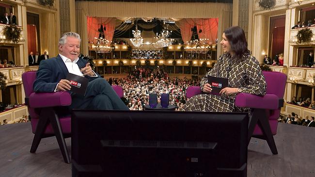 Christian Reichhold und Marion Nachtwey sitzen in violetten Armsesseln vor einer großen, festlich beleuchtetenOpernballkulisse, die den voll besetzten Saal zeigt.