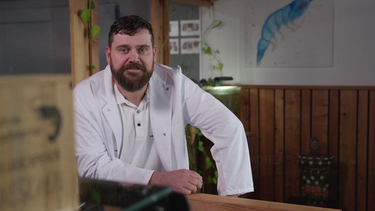 © Satel/ORF Andreas Aigner lehnt an einem Holztresen in einem Raum mit Holzwand und Aquarien im Hintergrund