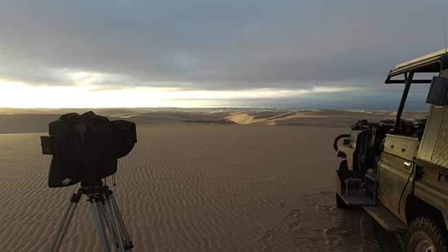 Ein Auto und eine Kamera stehen auf einer Sanddüne mit Blickrichtung über die Wüste. 