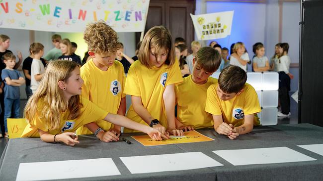 Fünf Volksschulkinder in gelben T-Shirts grübeln über ein Spiel.