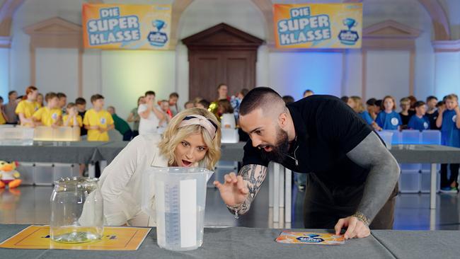 Die Moderator:innen Fanny und Stefan untersuchen einen gefüllten Messbecher. 