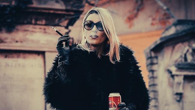 Die Kabarettistin Malarina trägt eine Sonnenbrille, raucht eine Zigarette und hält eine Bierdose in der Hand. Sie trägt schwarze Handschuhe und eine schwarze Kunstpelzjacke