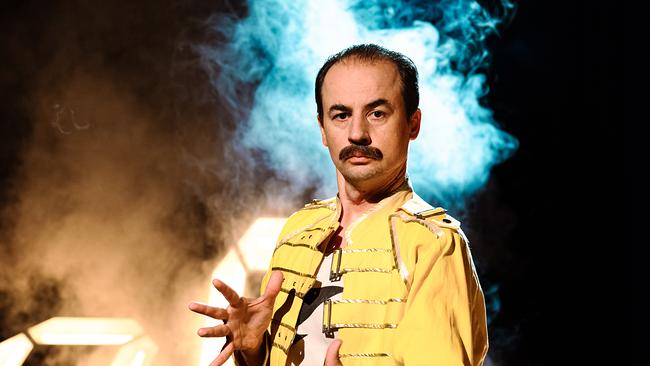 Austrofred in einem gelben Bühnenoutfit mit Schnurrbart, das an Freddie Mercury erinnert. Im Hintergrund sind farbige Nebeleffekte und dramatische Beleuchtung zu sehen.