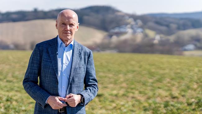 Peter Resetarits steht in blauem Sakko in einer ländlichen, hügeligen Landschaft mit Wiesen und einem Dorf im Hintergrund.