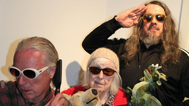 Drei Personen stehen vor einer weißen Wand: Links DJ Hell mit weißer Sonnenbrille und Bürste in der Hand, in der Mitte eine ältere Frau, Brigitte Meese, mit Mütze, Sonnenbrille und einem großen Stofftier im Arm, rechts Jonathan Meese mit langen Haaren, Sonnenbrille und Plüschdrachen, der salutiert. Alle drei blicken ernst oder entschlossen und tragen verspielte Requisiten.