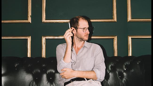 Wanda-Frontman und Autor Marco Wanda sitzt auf einem schwarzen Ledersofa vor einer dunkelgrünen Wand mit goldenen Rahmenleisten und hält eine Zigarette in der Hand, während er nach rechts blickt.
