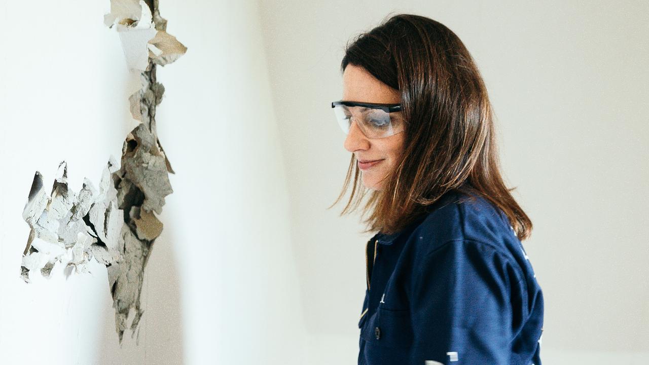 Lisa Gadenstätter schlägt mit Blaumann und Schutzbrille ausgestattet mit einem Hammer gegen die abbruchreife Wand.