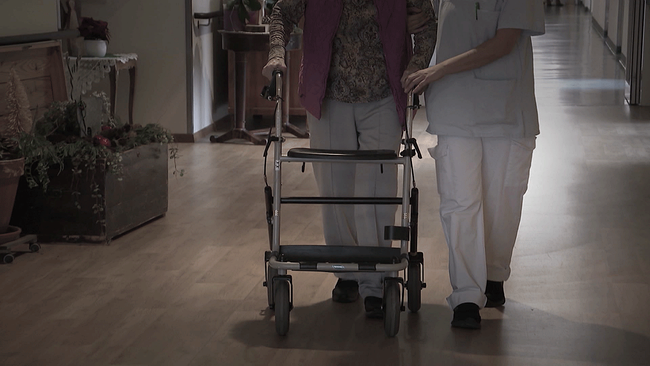 Person geht an Rollator und wird von Pflegekraft gestützt.