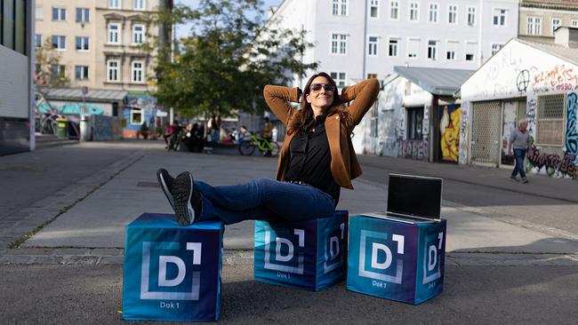 Lisa Gadenstätter sitzt auf Dok1-gebrandeten Sitzwürfeln auf einem öffentlichen Platz in Wien. Sie eröffnet einem "Dok1 Open Mic Corner": Hier bittet sie PassantInnen zum Gespräch und fragt nach, was die ÖsterreicherInnen von neuen Arbeitszeitmodellen halten. 