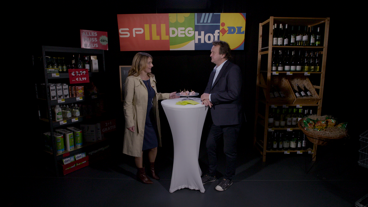 Hanno Settele steht neben Wirtschaftspsychologin Julia Pitters in einem eigens gebauten Supermarkt und bespricht mit ihr die gängigsten Verkaufstricks. Die beiden stehen einander an einem Stehtisch gegenüber. 