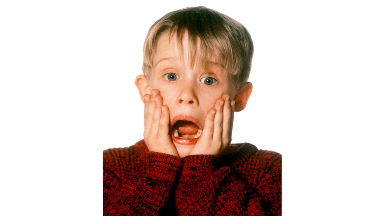 Macaulay Culkin als Kevin mit kurzem, blonden Haar in rotem Strickpullover vor weißem Hintergrund, erschrocken hält er beide Hände an seine Wangen