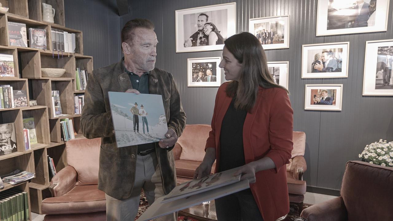 Anrold Schwarzenegger steht neben Lisa Gadenstätter und hält ihr das Bild vn einem Ausflug auf den Dachsteingletscher entgegen, welches sie ihm bei der Eröffnung des AWS Solution Hubs geschenkt hat