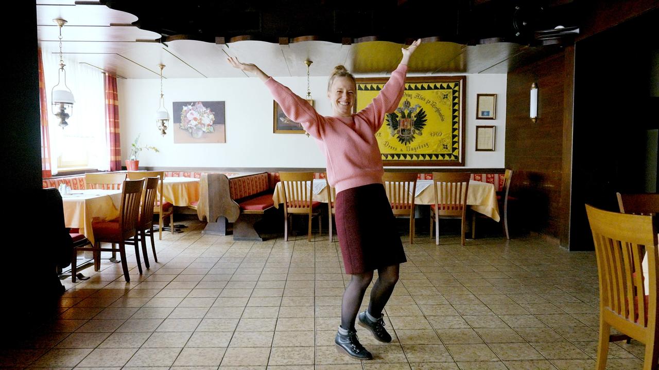 © Jenseide Kira Saskia Schinko ist zu Gast in Dross im Waldviertel, NÖ und steht in Pullover und Rock mit ausgestreckten Armen in einem leeren Gasthaus.