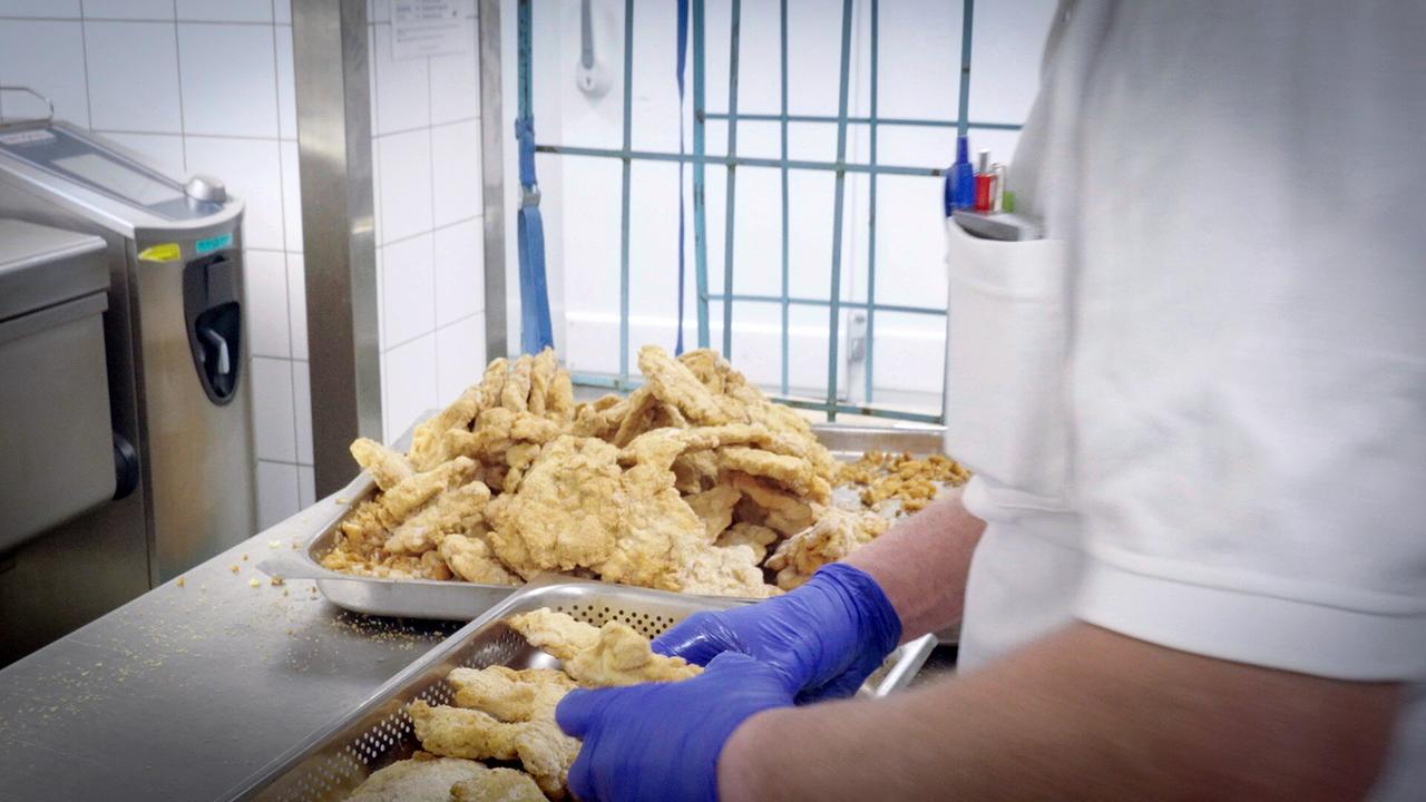 © ORF/Langbein & Partner Ein Mann mit blauen Handschuhen sortiert gefrorene Schnitzel von einem Tablett in ein Frittierblech mit Löchern