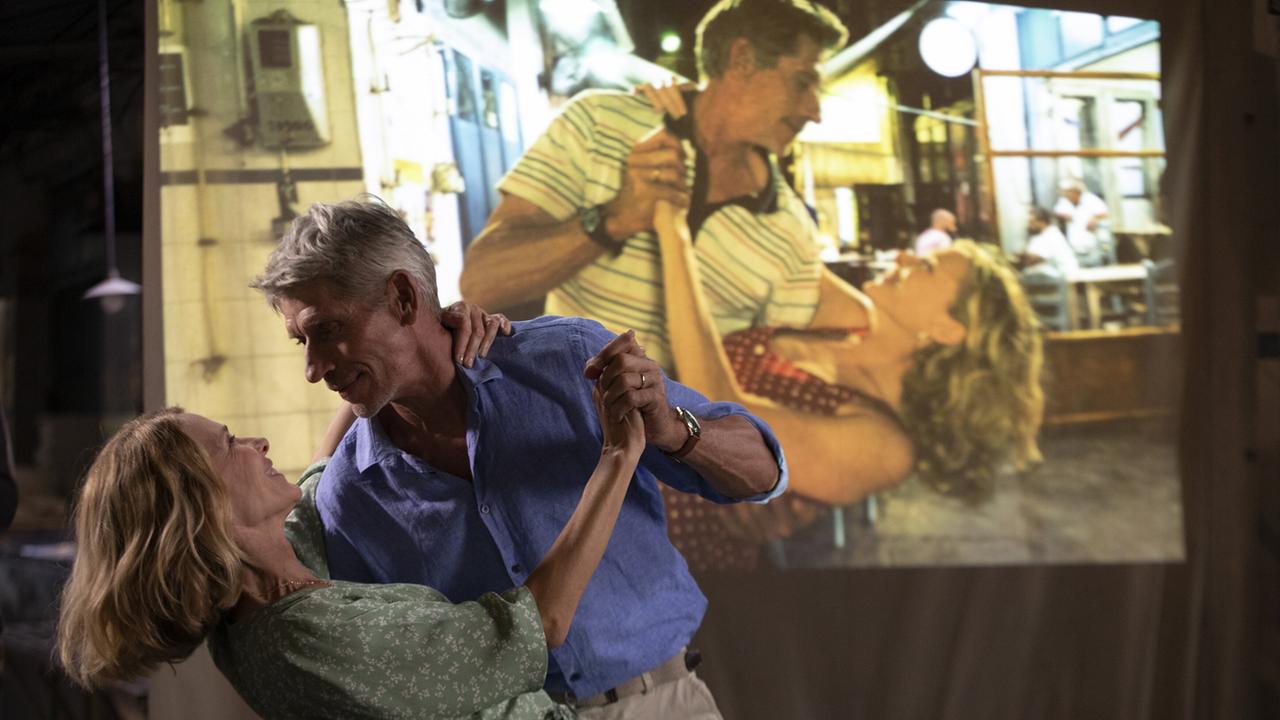 Claire und Thierry in einer Tanzpos, vor einer Leinwand, auf der ein Dia projiziiert wird, das die beiden auf einem alten Urlaubsfoto in derselben Pose zeigt