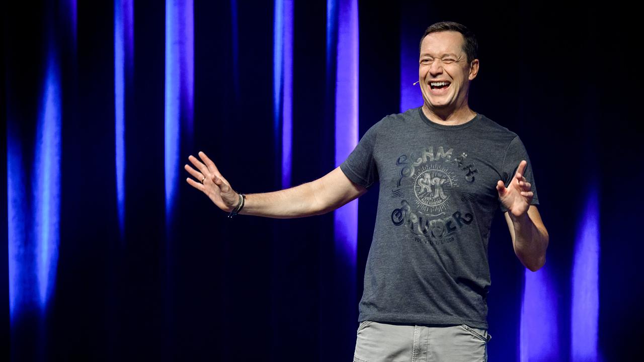 © ORF/Mutterschifffilm GmbH/Ulrike Rauch Alex Kristan steht auf einer Bühne vor einem blau beleuchtetem Vorhang und gestikuliert während der Aufführung.