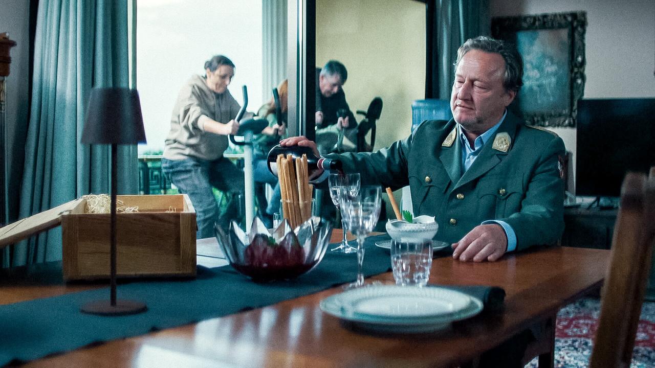 © ORF/Allegro Film Franz in grüner Uniform schenkt an einem gedeckten Esstisch Wein ein, während im Hintergrund trainiert wird.