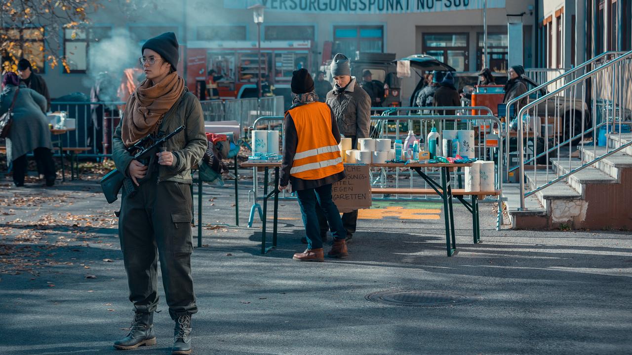 © ORF/Allegro Film/Anjeza Cikopano Vor einem Versorgungspunkt bewacht eine bewaffnete Frau einen belebten Platz mit Hilfsgütern und Einsatzkräften.