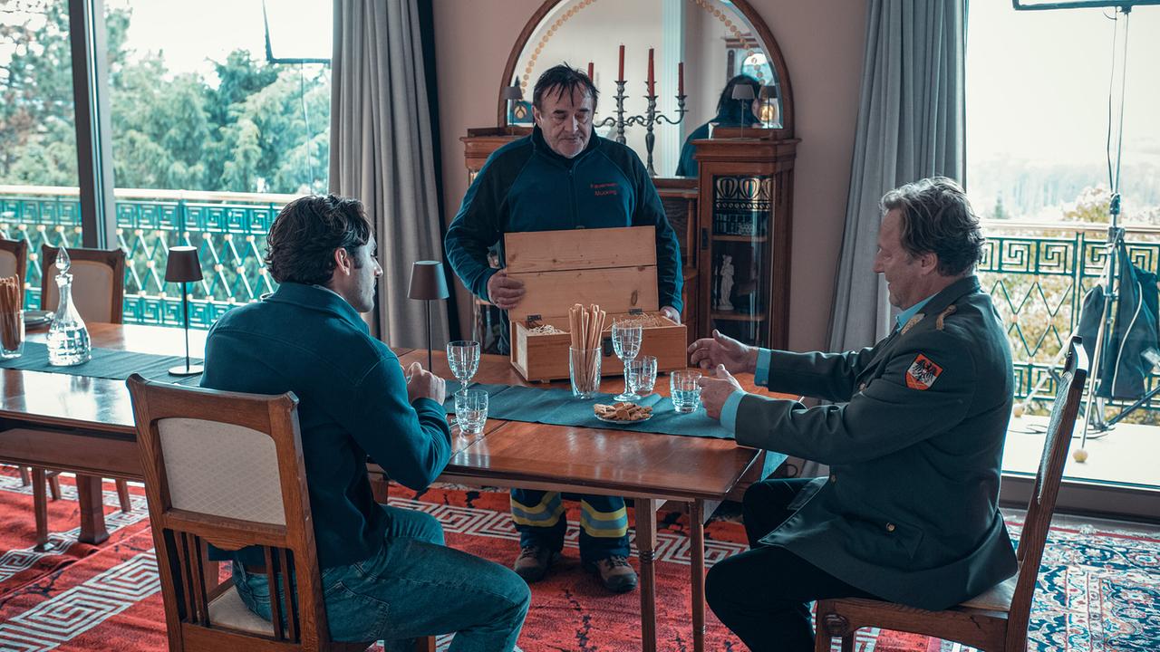 © ORF/Allegro Film/Stefan Robitsch Ein Mann präsentiert Georg und Franz, beide in grüner Uniform, an einem langen Esstisch den Inhalt einer Holzkiste.