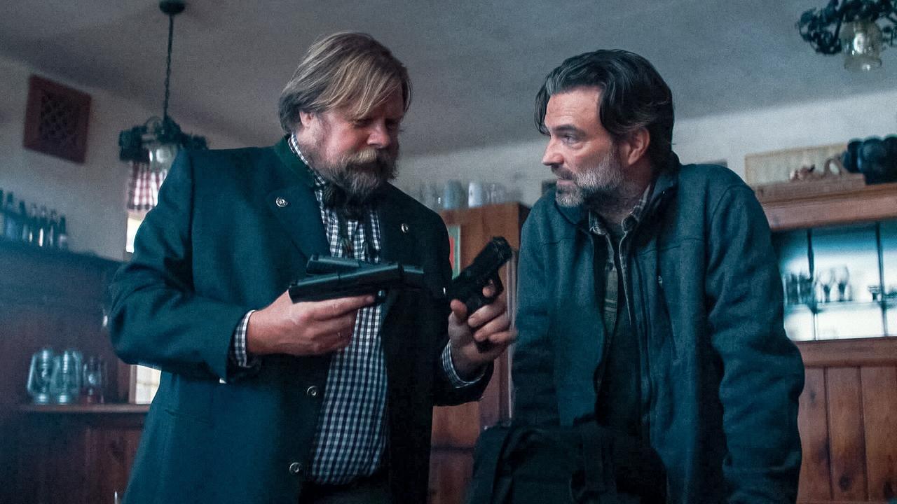 © ORF/Allegro Film In einem Gasthaus zeigt der bärtiger Jens seinem skeptischen Gegenüber Bernhard drei Pistolen, die er in Händen hält.