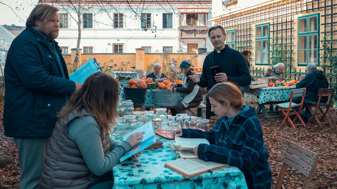 © ORF/Allegro Film/Anjeza Cikopano In einem herbstlichen Innenhof arbeiten mehrere Menschen, unter ihnen Carola und Agnes, an Tischen mit Kürbissen und Einmachgläsern, während Priester Matias und Jens mit Klemmbrett dabeistehen.