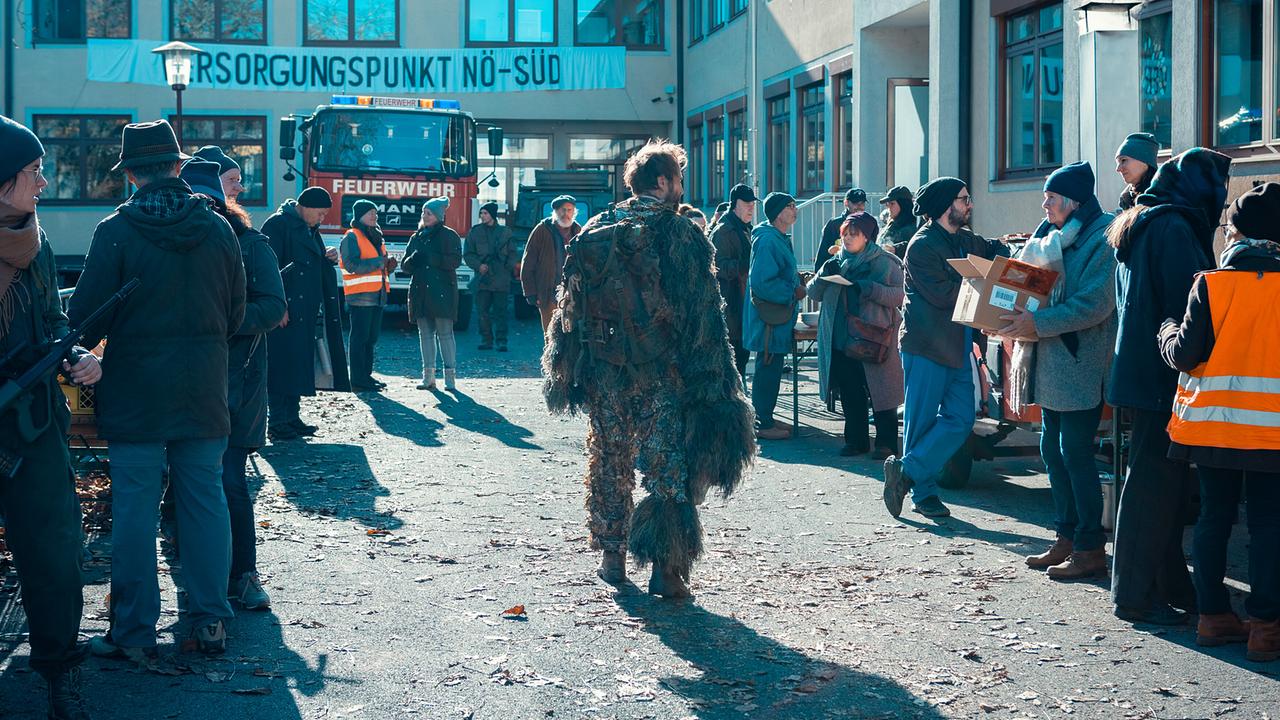© ORF/Allegro Film/Anjeza Cikopano Norbert in Tarnkleidung steht vor einem Gebäude mit der Aufschrift „Versorgungspunkt NÖ-Süd“, wo sich Menschen und Einsatzkräfte versammelt haben.