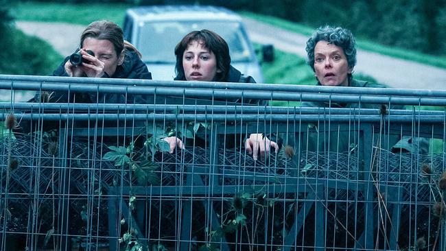 Agnes, Marie und Betti spähen über ein Absperrgitter im Grünen, während im Hintergrund ein Auto parkt.