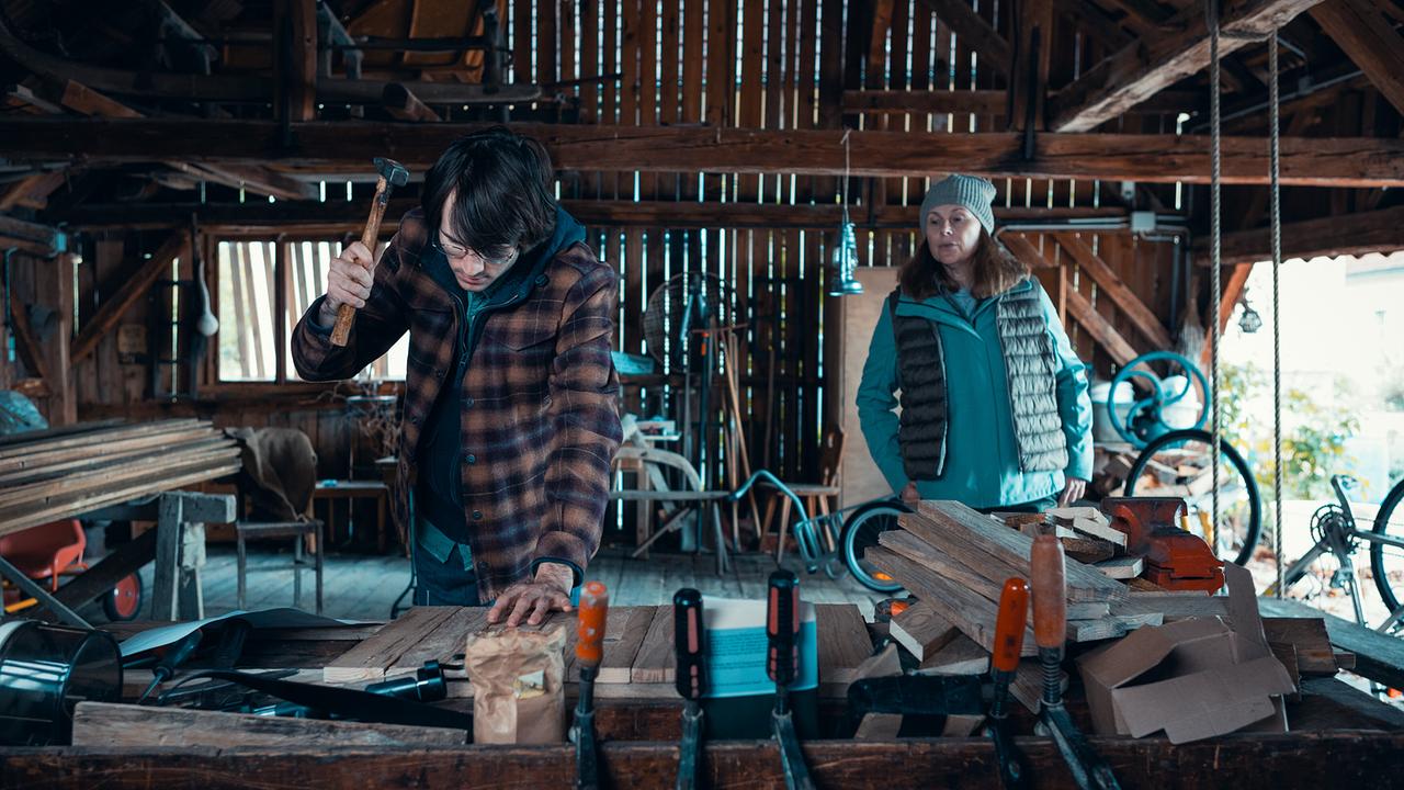 © ORF/Allegro Film/Anjeza Cikopano Jakob bearbeitet in einer Werkstatt Holz mit einem Hammer, während Carola zusieht.