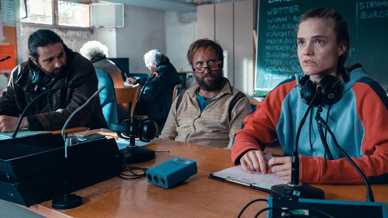 © ORF/Allegro Film/Anjeza Cikopano Laura mit Kopfhörern sitzt mit Norbert und weiteren Freiwilligien an einem Tisch mit Funkgeräten und Mikrofonen in einem Raum mit einer Schultafel im Hintergrund.