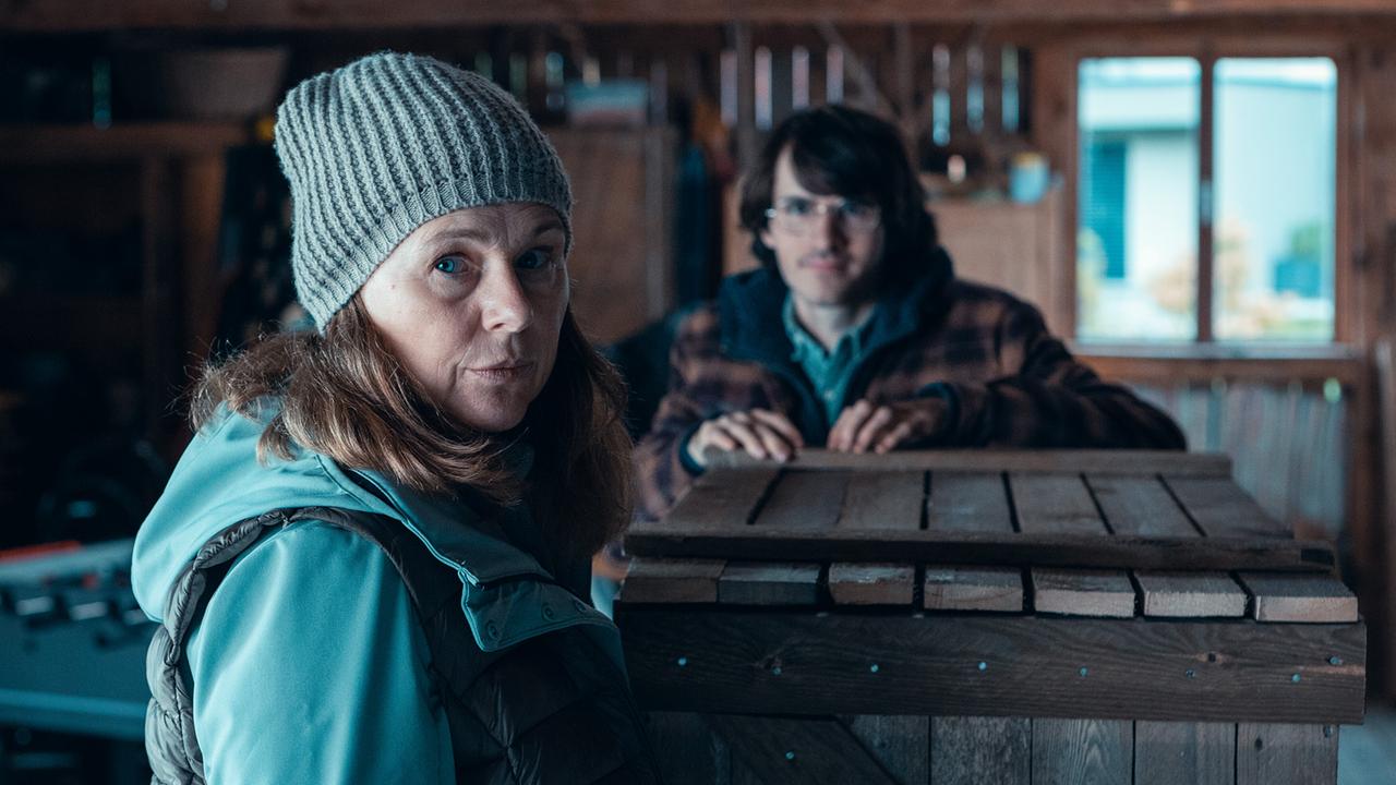 © ORF/Allegro Film/Anjeza Cikopano Carola mit grauer Strickmütze und Outdoorjacke blickt in einer rustikalen Werkstatt zur Kamera, während im Hintergrund Jakob hinter einer Holzkiste steht.