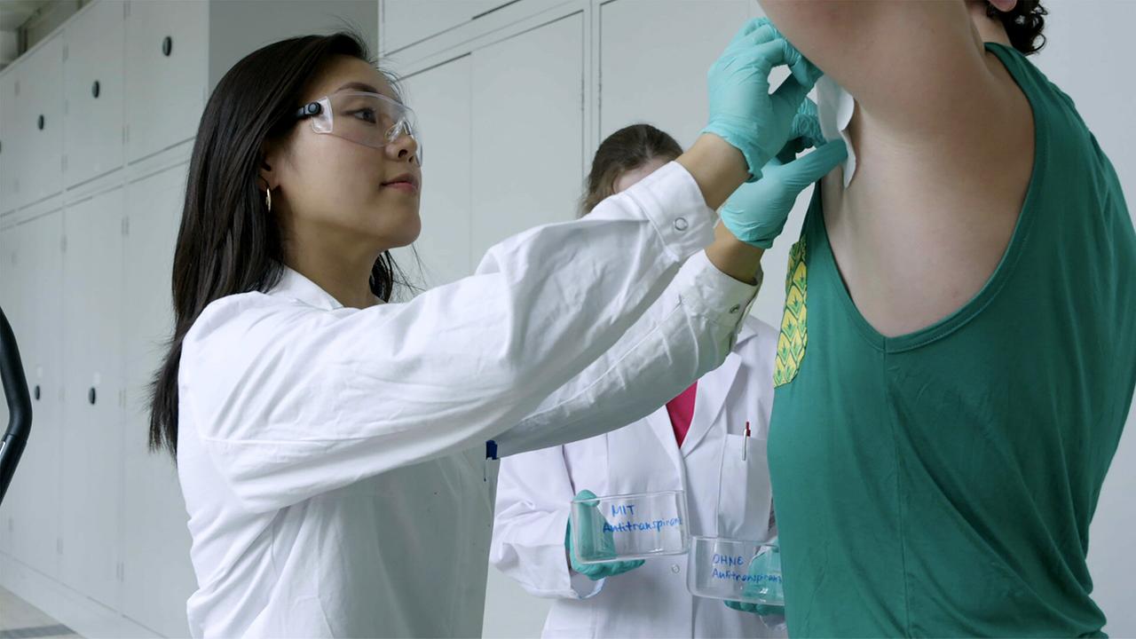 Eine junge Frau in weißem Laborkittel mit Schutzbrille und türkisen Handschuhen nimmt von der Achsel eines Probanden mit einem Tuch eine Schweißprobe