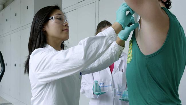 Eine junge Frau in weißem Laborkittel mit Schutzbrille und türkisen Handschuhen nimmt von der Achsel eines Probanden mit einem Tuch eine Schweißprobe