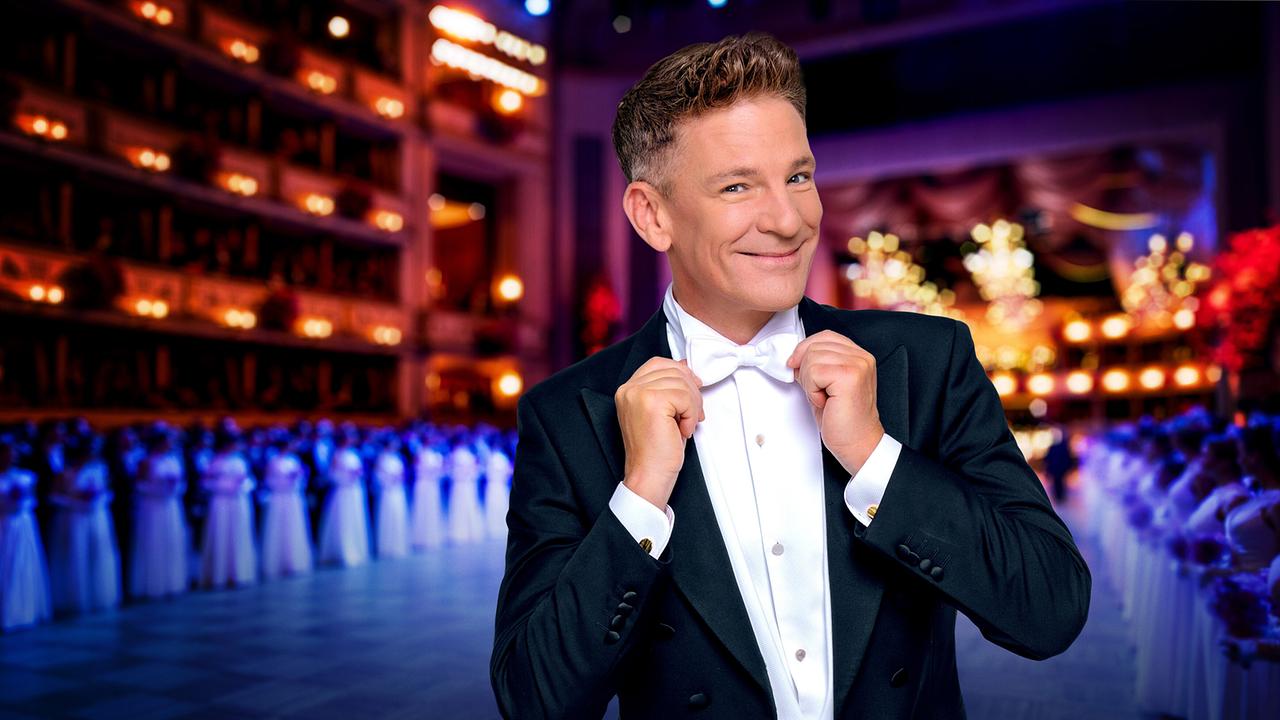 Andi Knoll im Frack richtet seine weiße Fliege, während im Hintergund der Ballsaal der Wiener Staatsoper mit den Debütantinnen und Debütanten des Wiener Opernballs zu sehen ist.