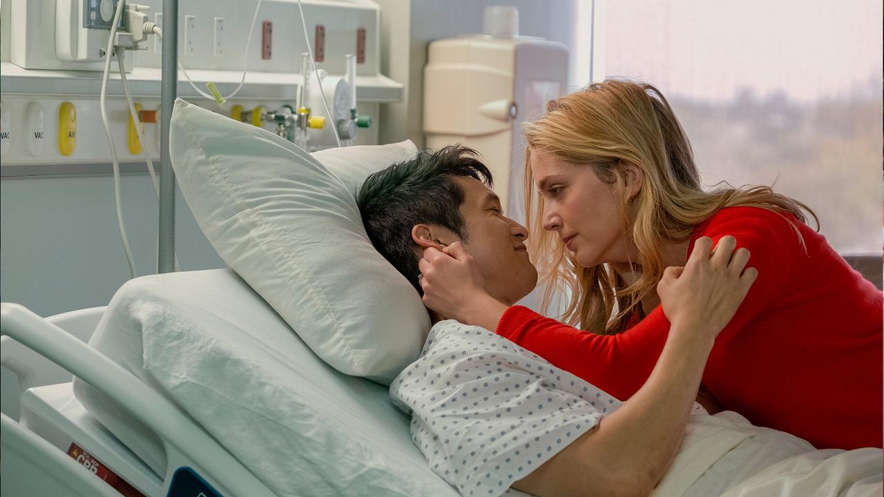 © ORF/Universal Solomon Chau liegt im Krankenbett im Spital, Jenn Carter beugt sich zu ihm und streichelt ihm am Ohr, lächelnd schaeun sich die beiden tief in die Augen