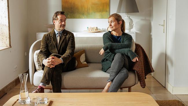 © ORF/LEONINE Studio/Wiedemann & Berg Film/Jürgen Olczyk Jan Josef Liefers als Heinz Hellmich im olivgrünen Schnürlsamt-Anzug und Nadja Uhl als Carla Hellmich in grauer Jeans, blauem Shirt und grüner Strickjacke, auf einer Couch sitzen sie mit Abstand nebeneinander und blicken sich freundlich von der Seite an