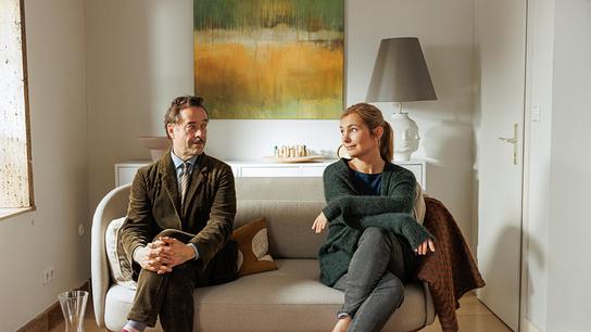 Jan Josef Liefers als Heinz Hellmich im olivgrünen Schnürlsamt-Anzug und Nadja Uhl als Carla Hellmich in grauer Jeans, blauem Shirt und grüner Strickjacke, auf einer Couch sitzen sie mit Abstand nebeneinander und blicken sich freundlich von der Seite an 