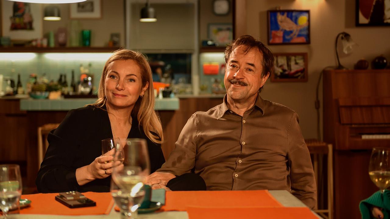© ORF/LEONINE Studio/Wiedemann & Berg Film/Jürgen Olczyk Nadja Uhl als Carla Hellmich und Jan Josef Liefers als Heinz Hellmich sitzen lächelnd an ihrem Wohnzimmertisch und unterhalten sich freundlich mit ihren Gästen
