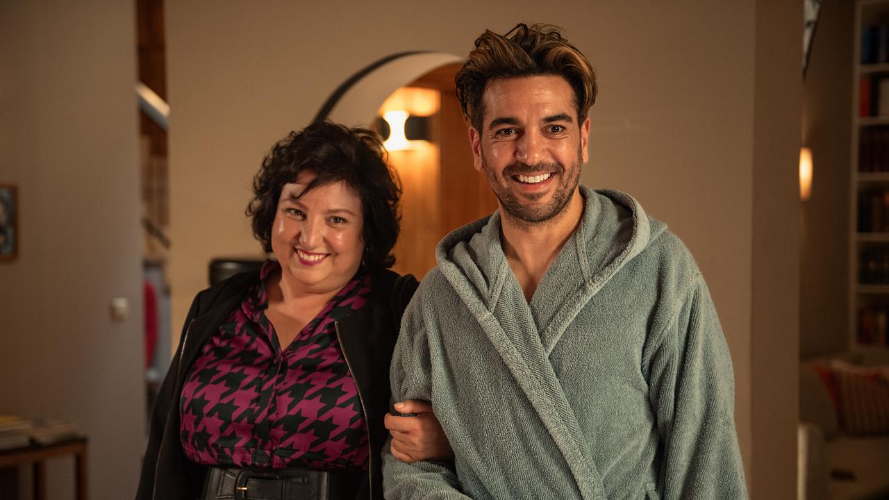 © ORF/LEONINE Studio/Wiedemann & Berg Film/Jürgen Olczyk Meltem Kaptan als Kiraz Tüfek im lila-schwarzem Hahnentritt-Kleid und Elyas M’Barek als Älex Sahavi im grauen Bademantel, die beiden stehen im Wohnzimmer der Hellmcihs und lächeln freundlich ihr Gegenüber an
